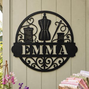 May include: A circular black metal wall sign featuring a central dress form, a spool of thread, and a needle. The design includes decorative scrollwork and the name EMMA in bold capital letters centred within a horizontal rectangular banner.