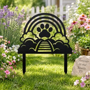 May include: A black metal garden stake featuring a cutout design of a paw print centered above a path leading into clouds, framed by a rainbow arch. The silhouette includes two pointed prongs at the base for insertion into the ground.