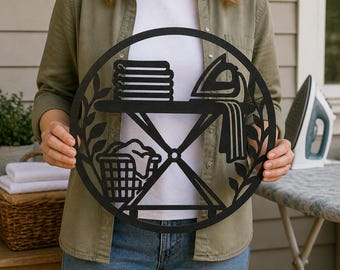 May include: A circular black metal wall sign featuring a cutout design of laundry room items. The graphic includes a stack of folded towels, an iron, an ironing board, and a laundry basket, all framed by a decorative leaf pattern border.