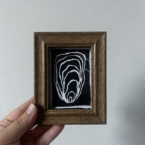 Könnte beinhalten: Ein kleiner rechteckiger Holzrahmen mit einem schwarz-weißen Druck. Das Kunstwerk zeigt eine Reihe ineinander verschachtelter, organischer weißer Linien auf einem soliden schwarzen Hintergrund, die an den Querschnitt einer Auster erinnern.