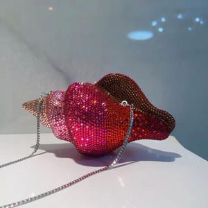 Könnte beinhalten: Eine muschelförmige Clutch, die vollständig mit funkelnden Kristallen bedeckt ist, die von hellem Rosa und Pfirsich an der Spitze zu leuchtendem Magenta, tiefem Rot und dunkler Bronze übergehen. Sie hat einen dünnen, silberfarbenen Metallkettenriemen.