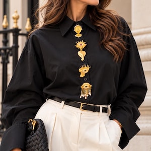 May include: A black long-sleeved button-down shirt featuring a vertical row of five distinct gold-toned decorative brooches. The shirt is tucked into cream-coloured trousers with a thin black leather belt and paired with a black woven clutch bag.