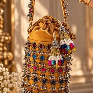May include: A mustard yellow bucket bag featuring a drawstring closure and a handle adorned with silver-toned metal flower charms. The exterior is decorated with rows of silver flower motifs containing colorful inlays and dangling silver beads.