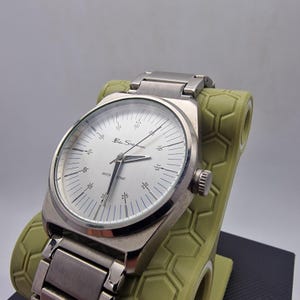 A authentic pre-owned Ben Sherman BS095 Men's Quartz Watch. Featuring a unique brushed silver analogue display, this timepiece stands out with its striking blue-accented numerical markers and precise 3-hand design. The durable stainless steel case is paired with a matching silver-toned metal link bracelet,