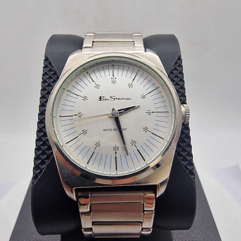 A authentic pre-owned Ben Sherman BS095 Men's Quartz Watch. Featuring a unique brushed silver analogue display, this timepiece stands out with its striking blue-accented numerical markers and precise 3-hand design. The durable stainless steel case is paired with a matching silver-toned metal link bracelet,