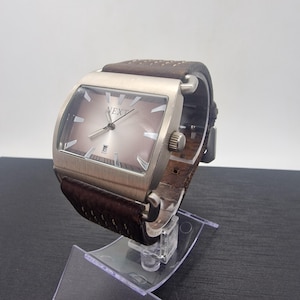 Puede incluir: Un reloj de pulsera rectangular con caja de metal plateado cepillado y correa de cuero marrón con costuras decorativas. La esfera es de color marrón degradado con marcadores de hora plateados, manecillas plateadas y el texto NEXT arriba.