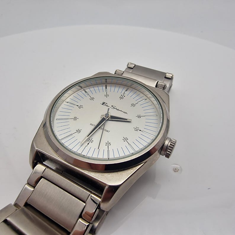 A authentic pre-owned Ben Sherman BS095 Men's Quartz Watch. Featuring a unique brushed silver analogue display, this timepiece stands out with its striking blue-accented numerical markers and precise 3-hand design. The durable stainless steel case is paired with a matching silver-toned metal link bracelet,