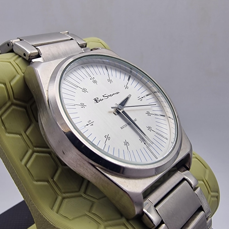 A authentic pre-owned Ben Sherman BS095 Men's Quartz Watch. Featuring a unique brushed silver analogue display, this timepiece stands out with its striking blue-accented numerical markers and precise 3-hand design. The durable stainless steel case is paired with a matching silver-toned metal link bracelet,