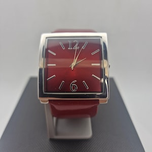 Könnte beinhalten: Eine quadratische Armbanduhr mit einem tiefroten Zifferblatt, silberfarbenen Stundenmarkierungen und Zeigern. Die Uhr hat ein poliertes silberfarbenes Metallgehäuse und ein passendes rotes Armband. Die Zahlen 12 und 6 sind in Silber abgebildet.