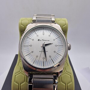 A authentic pre-owned Ben Sherman BS095 Men's Quartz Watch. Featuring a unique brushed silver analogue display, this timepiece stands out with its striking blue-accented numerical markers and precise 3-hand design. The durable stainless steel case is paired with a matching silver-toned metal link bracelet,
