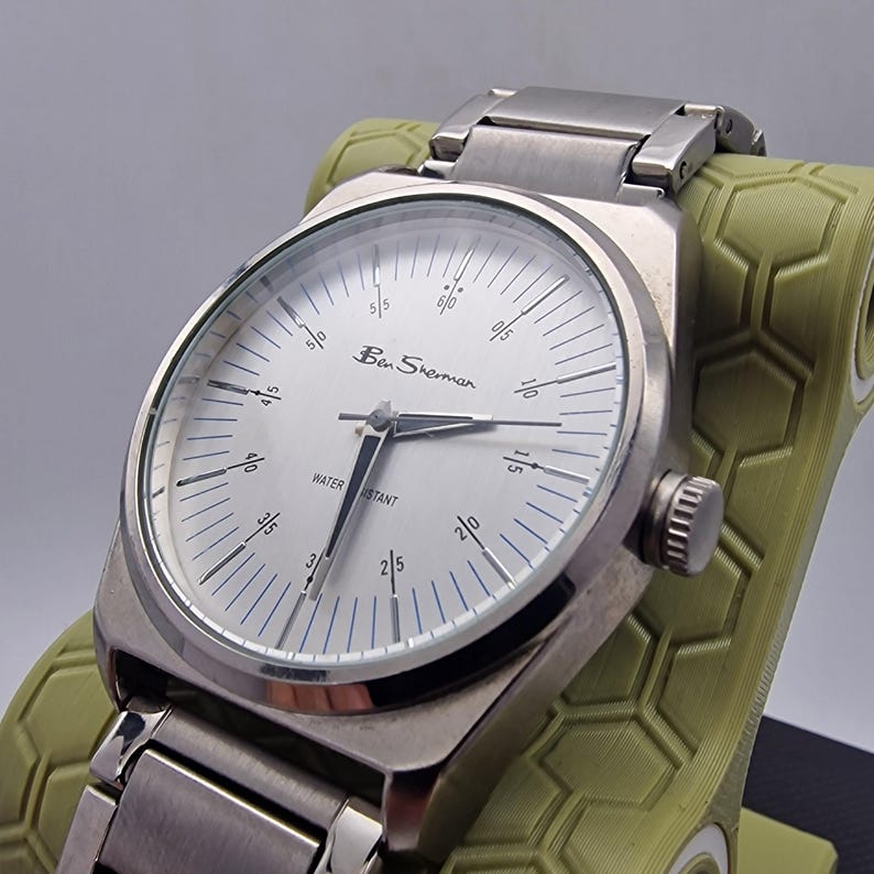 A authentic pre-owned Ben Sherman BS095 Men's Quartz Watch. Featuring a unique brushed silver analogue display, this timepiece stands out with its striking blue-accented numerical markers and precise 3-hand design. The durable stainless steel case is paired with a matching silver-toned metal link bracelet,
