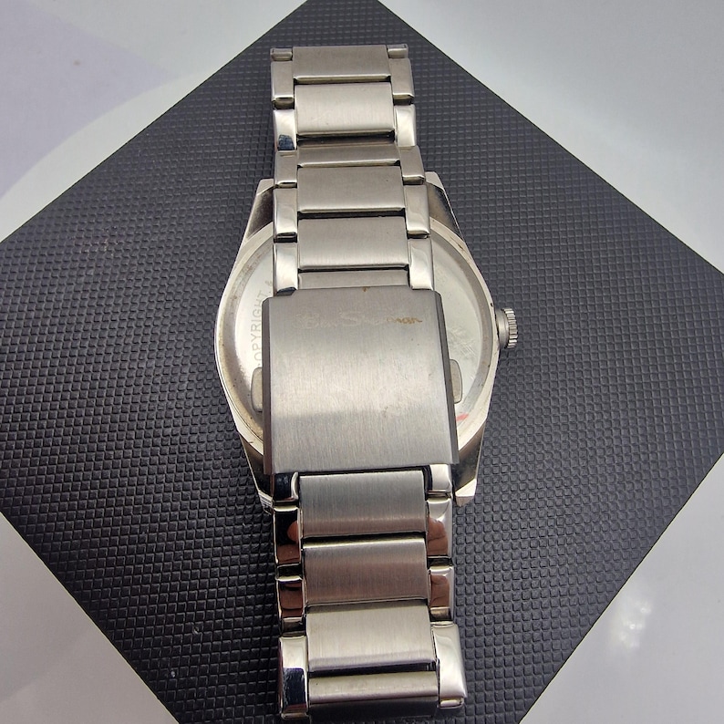 A authentic pre-owned Ben Sherman BS095 Men's Quartz Watch. Featuring a unique brushed silver analogue display, this timepiece stands out with its striking blue-accented numerical markers and precise 3-hand design. The durable stainless steel case is paired with a matching silver-toned metal link bracelet,