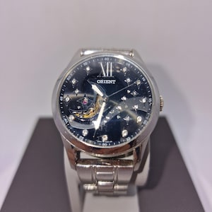 Peut inclure: Une montre-bracelet Orient argentée avec un cadran noir orné d'un motif d'étoiles, d'index en cristal et d'une fenêtre révélant le mouvement mécanique. Elle possède un bracelet à maillons en acier inoxydable et un boîtier rond avec couronne.