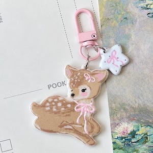 May include: A keyring featuring a tan fawn with white spots, a pink bow around its neck, and a small pink bow on its ear. The charm is attached to a light pink lobster clasp and includes a small white star-shaped charm with a pink bow design.