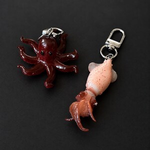 May include: Two keychains featuring marine life figures. One is a dark reddish-brown octopus with eight tentacles and black eyes. The other is a pale pink and orange squid with speckled markings, multiple tentacles, and a silver metal lobster clasp.