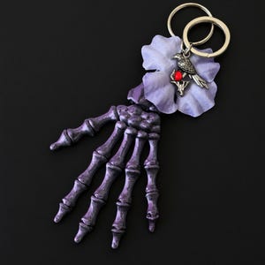 May include: A keychain featuring a purple skeleton hand with intricate etched details, attached to a light purple flower petal. A silver-toned bird charm with a small, bright red circular accent is positioned on the flower, connected to two silver metal rings.