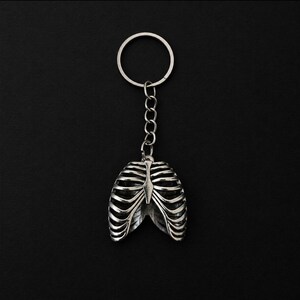 May include: A silver-toned metal keychain featuring a three-dimensional anatomical rib cage pendant. The pendant is attached to a standard circular split ring by a short metal chain. The design displays detailed horizontal rib bones.
