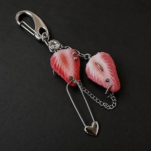 May include: A silver-toned metal keyring featuring a lobster clasp, a small clear rhinestone, and two textured red strawberry slices. The strawberry slices are connected by silver chains and a large safety pin charm with a small heart-shaped end.