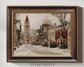 May include: A framed wall art print depicting a snowy European village street during winter. The scene features a tall clock tower with a red roof, half-timbered buildings, and red brick structures decorated with glowing string lights and Christmas trees.