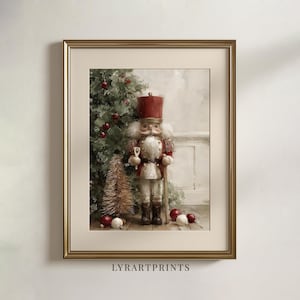 Peut inclure: Une impression d'art encadrée représentant un soldat casse-noisette en uniforme rouge et or, debout à côté d'un sapin de Noël vert, d'un petit sapin doré en écouvillon et d'ornements rouges et blancs dispersés sur un sol clair.