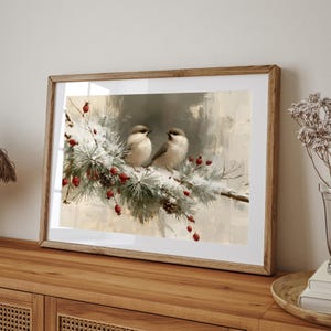 May include: A framed wall art print featuring two small birds perched on a snow-covered pine branch with red berries. The artwork displays a soft, painterly style with muted beige and white tones, encased in a light brown wooden frame.