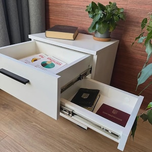 Könnte beinhalten: Ein weißer Nachttisch mit zwei offenen Schubladen und schwarzen Griffen. Die obere Schublade enthält ein englisches Buch. Die untere Schublade enthält einen dunklen Reisepass und ein kastanienbraunes Dokument mit goldenem Emblem. Obenauf liegen ein Buch und eine grüne Pflanze.