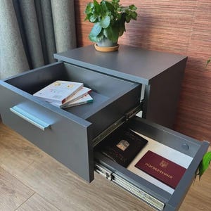 Op de afbeelding: Een donkergrijs zwevend nachtkastje met een open lade met zilveren handgreep waarin twee kleine boeken liggen. Een tweede onderste lade is uitgeschoven en bevat een zwart paspoort en een bordeauxrood document met de tekst POSVIDCHENNYA.