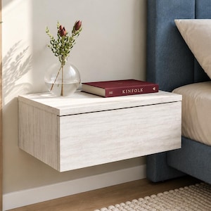 May include: A wall-mounted floating nightstand with a light wood grain finish. On top sits a clear glass vase containing green stems with small dark red buds, and a maroon hardcover book with the word KINFOLK printed in gold on the spine.