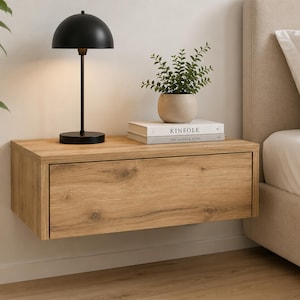 May include: A wall-mounted floating bedside table crafted from light oak wood with a single drawer. On top sits a black dome-shaped table lamp and a small potted green plant resting on a white book titled KINFOLK with the subtitle THE KINFOLK HOME.