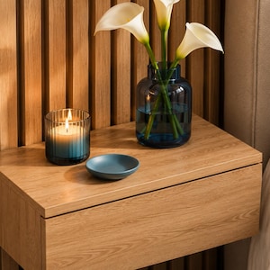 May include: A floating light wood nightstand with a drawer, featuring a blue glass vase with three white calla lilies, a small blue ceramic dish, and a ribbed glass candle holder containing a lit candle with a blue and white wax gradient.