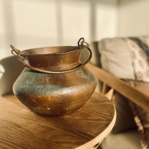 May include: A round, hammered copper pot with a wide opening and a curved metal handle attached to two side loops. The exterior surface displays a weathered, brownish-orange patina with visible areas of green oxidation and a textured finish.