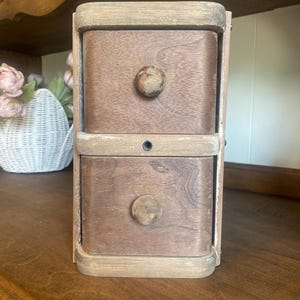 May include: A small wooden storage unit featuring two stacked drawers with rounded edges and circular wooden knobs. The unit has a weathered, light-toned frame with darker brown drawer fronts and a single small metal hole centered between them.