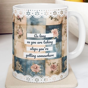 May include: A white ceramic mug featuring a patchwork pattern of blue and beige squares with floral motifs. The top and bottom edges have a white lace-like trim. The text reads: As long as you are taking steps you're getting somewhere.