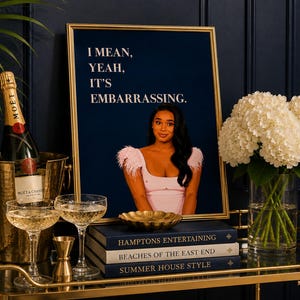 May include: A framed print featuring a person in a light pink top with feathered sleeves against a dark blue background, with the text "I MEAN, YEAH, IT'S EMBARRASSING." The print is displayed alongside a champagne bottle, glasses, and books.