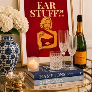 May include: A framed art print featuring a blonde woman in a red dress with the text EAR STUFF?!, displayed on a gold bar cart with a blue and white vase, a lit candle, two crystal champagne flutes, a crystal tumbler, and a bottle of Veuve Clicquot.