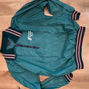 May include: A teal nylon pullover windbreaker jacket featuring a quarter-zip front, a collar, and long sleeves. The garment has dark green ribbed cuffs and hem with thin white stripes. The text "Americana Lake Geneva Resort" is printed in white.