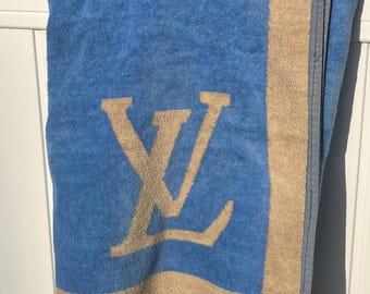 May include: A blue and beige terry cloth towel featuring a large, centered tan Louis Vuitton monogram logo. The towel has a wide beige border along the right side and bottom edge, finished with a dark grey stitched hem along the outer perimeter.