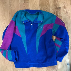 Może przedstawiać: Bluza polarowa z długim rękawem o wzorze typu color-block w kolorach kr&oacute;lewskiego błękitu, turkusu i magenty. Odzież posiada kołnierz zapinany na zatrzaski, ściągacz u dołu oraz wyhaftowane małe litery FILA w g&oacute;rnej części lewej strony klatki piersiowej.