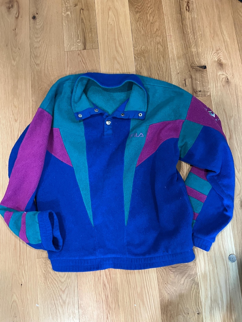 Może przedstawiać: Bluza polarowa z długim rękawem o wzorze typu color-block w kolorach kr&oacute;lewskiego błękitu, turkusu i magenty. Odzież posiada kołnierz zapinany na zatrzaski, ściągacz u dołu oraz wyhaftowane małe litery FILA w g&oacute;rnej części lewej strony klatki piersiowej.