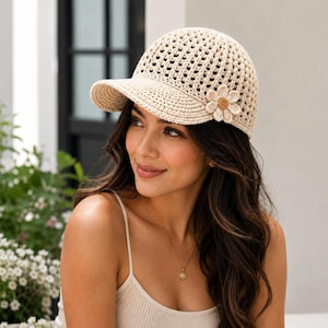 May include: A beige crochet baseball cap featuring an open-knit pattern and a stiff brim. The cap is adorned with a single, six-petaled fabric flower appliqué on the right side, which has a small, round, brown centre button detail.