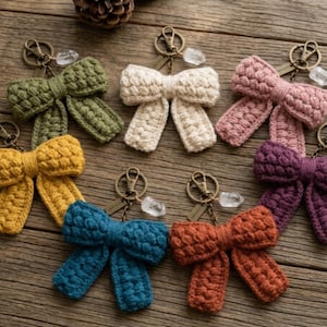 May include: Six crochet bow keychains in olive green, cream, dusty pink, mustard yellow, teal, and burnt orange. Each bow features a textured popcorn stitch pattern, a bronze-toned metal lobster clasp, and a small clear crystal charm attached to the ring.