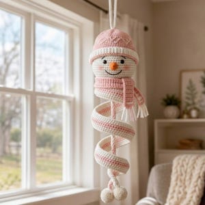 Könnte beinhalten: Ein hängendes Häkel-Schneemann-Ornament mit einer rosa-weißen Mütze, einem passenden rosa-weiß gestreiften Schal und einem spiralförmigen Körper. Der Schneemann hat eine kleine orangefarbene Nase, schwarze Augen, ein Lächeln und zwei Bommeln.