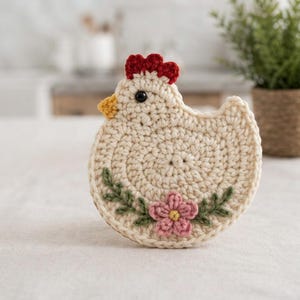 Puede incluir: Un posavasos de ganchillo color crema con forma de gallina, que presenta una cresta roja, un pico amarillo y un ojo negro. El cuerpo está decorado con una pequeña flor rosa con centro amarillo, flanqueada por dos ramas verdes bordadas.