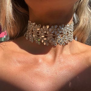 Puede incluir: Un collar tipo gargantilla ancho que presenta una disposición densa e intrincada de cristales transparentes de talla marquesa engastados en un marco de metal dorado. El diseño consta de múltiples formas de hojas superpuestas que envuelven el cuello, creando una apariencia texturizada y brillante con superficies reflectantes.