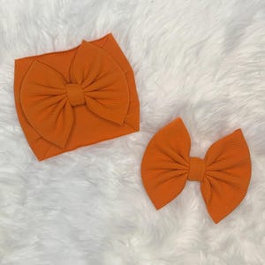 Könnte beinhalten: Ein leuchtend oranges Stoffstirnband mit einer großen, texturierten Schleife und eine passende separate Haarspange aus orangem Stoff. Beide Artikel bestehen aus einem weichen, gerippten Material mit einer durchgehend einheitlichen orangen Farbe.