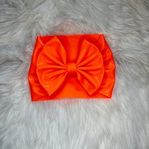 May include: A bright neon orange headband featuring a large, centred, multi-layered fabric bow. The bow is constructed with gathered, pleated fabric that creates a voluminous, fan-like appearance, securely attached to a wide, matching orange band.