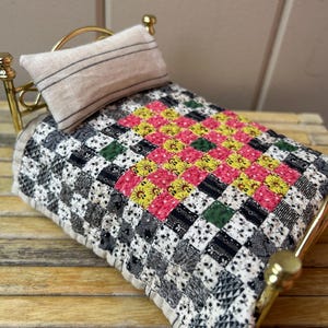 May include: A miniature brass bed frame featuring a beige pillow with thin black stripes and a patchwork quilt. The quilt displays a grid of small squares in black, white, yellow, pink, and green floral and geometric patterns.