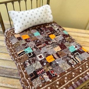 May include: A miniature metal bed frame with a brass finish, featuring a white rectangular pillow with a small grey dot pattern and a patchwork quilt. The quilt is composed of various brown, orange, and turquoise fabric squares.
