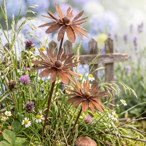 May include: Three rust-colored metal flower sculptures with pointed petals and spherical centers stand among green grass, wild strawberries, and two small mushroom figurines, one brown and one red with white spots, in a garden setting.