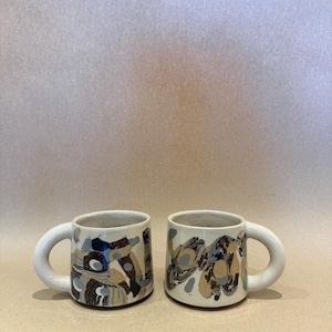 May include: Two ceramic mugs with a cream-colored base featuring an abstract pattern of dark brown, tan, and light blue shapes. Each mug has a thick, rounded handle and a cylindrical body with a smooth finish and a wide, circular opening.